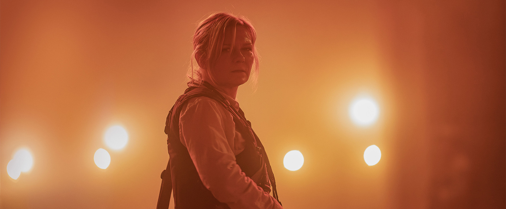 A woman wearing a bullet proof vest stood in the middle of a road with bright spotlights behind her. The air is filled with dust and has an orange hue to it.