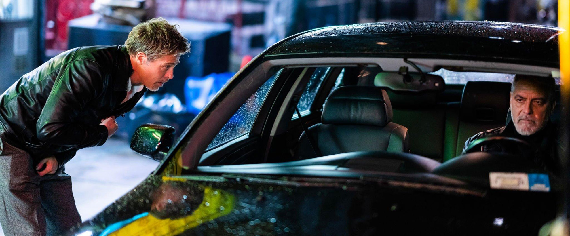 A man leaning down to talk to another man sat in a car, they are in a city in the rain.