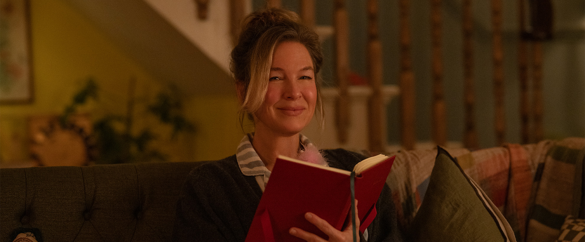 Still from Bridget Jones Made About the Boy. Bridget Jones sitting on sofa with a book