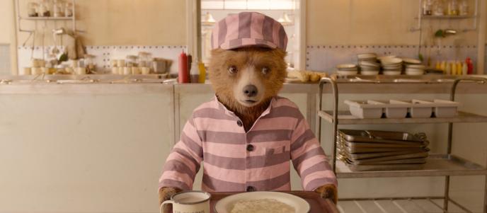 Paddington Bear wearing a pink striped prison uniform, holding a dinner tray in a kitchen.