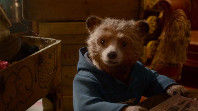 paddington bear wearing his blue botton up hooded coat sitting with his paws on the table. e is looking to the left