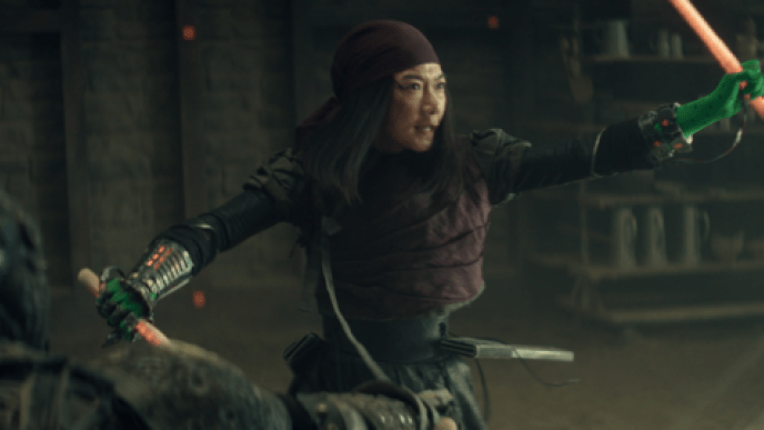 A woman with her arms covered in green screen material, holding plastic swords stand ins, they are clashing with other plastic swords coming from outside the frame. 