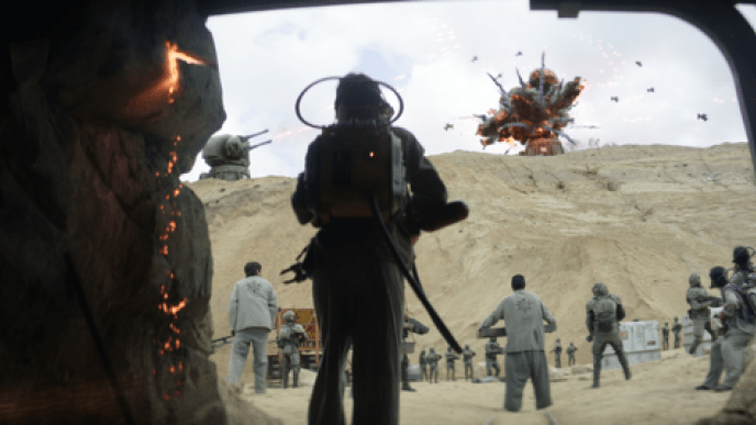 A group of people stood looking at an explosion happening at the top of a sand dune in the background, the people are holding guns. 