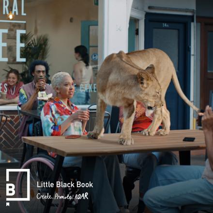 A still from an ITV advert showing a lioness standing on a cafe table surrounded by people who look shocked. 