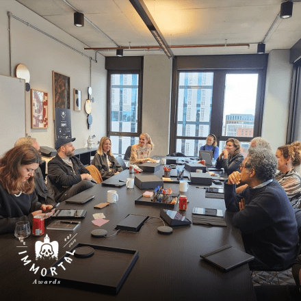  A photograph of the Immortal Awards judges sitting around a table viewing content on a screen. There is the Immortal Awards logo in the bottom left corner.